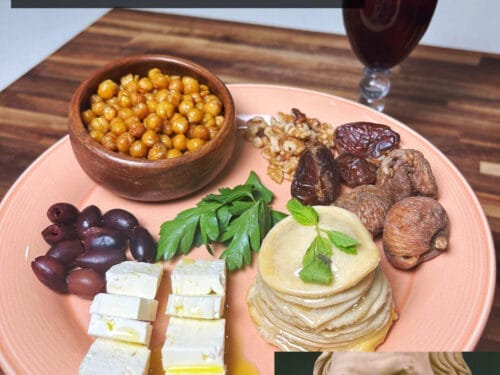 Wooden bowl of chickpeas, pink plate with cheese, olives, flatbread, roasted vegetables, and wine glass