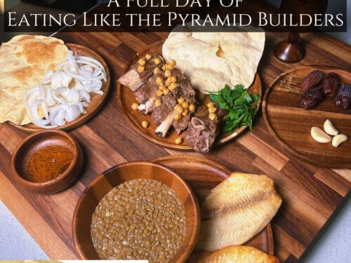 Wooden boards holding flatbread, cooked fish fillets, meat with chickpeas, sliced onions, fresh herbs, dates, garlic, grain porridge, and sauce in bowls