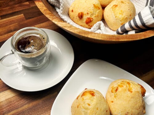 Golden-brown cheese breads served with dark coffee, wooden bowl, and white plate on wood table