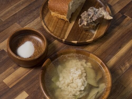 Wooden bowls containing bread, fish, potatoes with onions, and salt on wooden surface