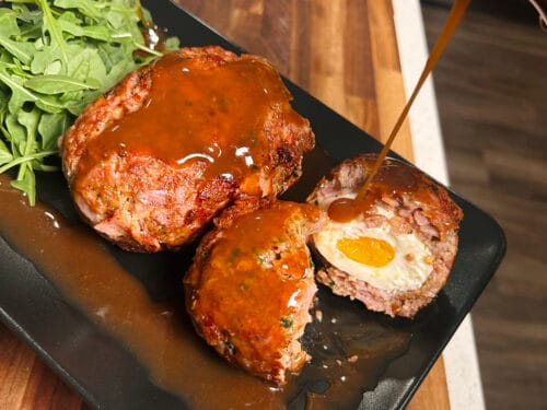 Breaded meatballs with egg centers glazed in brown sauce, served on black plate with arugula