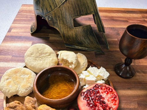 Metal helmet with red crest, wooden plate with flatbread, figs, cheese, olives, pomegranate half, oil bowl, and goblet on table