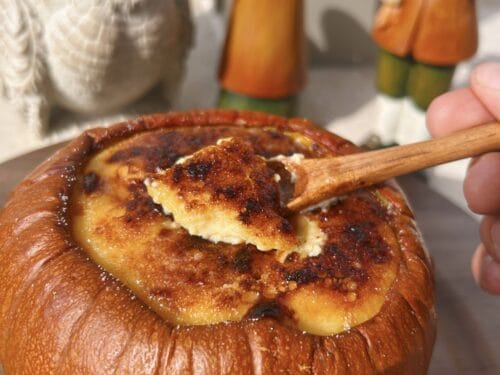 Wooden spoon lifting pale custard with browned top from hollowed whole pumpkin