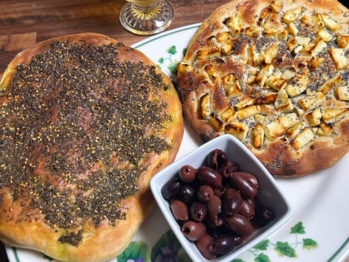 Two topped flatbreads and bowl of olives on decorative plate, with golden beverage in glass