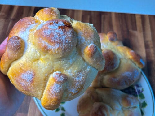 Hand holding a golden loaf dusted with sugar, with protruding rounded knobs, on a plate with similar loaves