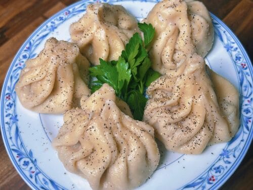 Steamed dumplings with crinkled edges coated in black pepper, centered with fresh parsley on blue-patterned plate