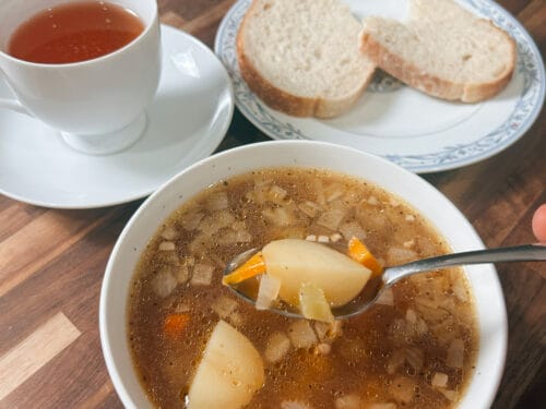 Spoon lifting potato and carrot from brown broth; cup of tea and two bread slices on wooden table
