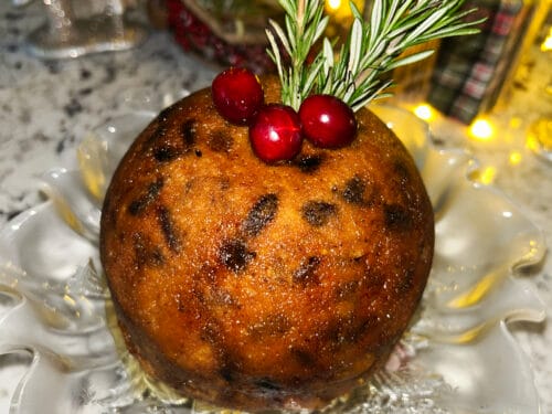 Round golden-brown cake with dark spots, topped with fresh rosemary and red baubles on white plate
