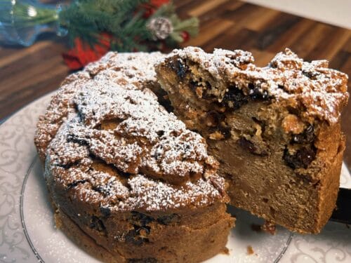 Whole spiced cake with slice removed, heavily dusted with powdered sugar, surrounded by lit candles and evergreen branches