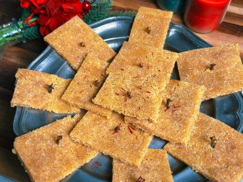 Tan rectangular spiced cake pieces with visible dark specks arranged in star pattern on dark plate, surrounded by red flowers, candles, and greenery