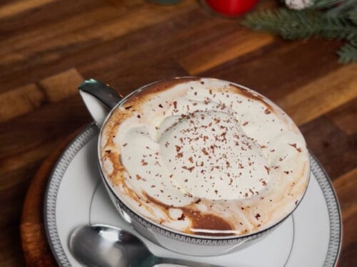 Foamy beverage topped with cocoa powder in white cup on wooden table, surrounded by lit candles and greenery
