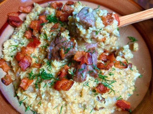 Creamy grain porridge topped with browned meat cubes and fresh chives in earthenware bowl