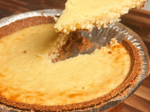 Pale yellow cheesecake with brown crust in aluminum tin, one slice lifted on server