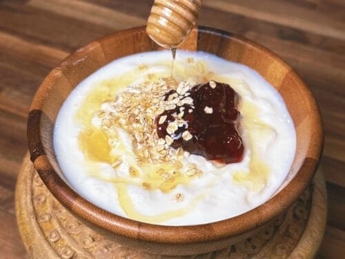 White yogurt in carved wooden bowl topped with oats, dark jam, honey drizzling from wooden dipper