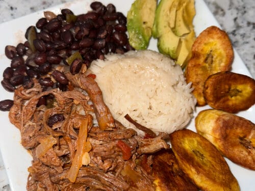 Plate with shredded beef, white rice, dark beans, golden fried plantain rounds, and avocado slices