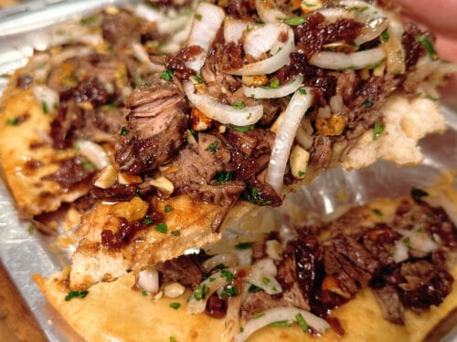 Hand holding a slice of flatbread piled with shredded braised meat, raw onion slices, and green herbs