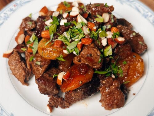 Brown meat pieces topped with orange dried fruit, sliced almonds, and green herbs on blue-patterned white plate