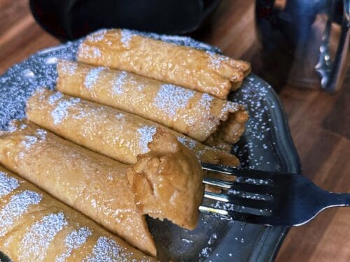 Six rolled pastries dusted with powdered sugar on dark plate with fork and skillet