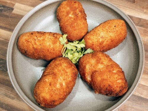 Five golden-brown oblong croquettes arranged in flower pattern on round gray plate with celery leaf garnish