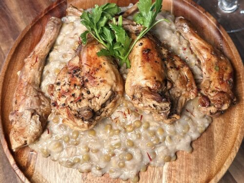 Roasted rabbit legs with pearl-colored pottage on wooden plate, topped with fresh parsley