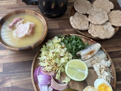 Wooden board with fish, pickles, hard biscuits, onion, greens, eggs, lime, and a bowl of broth with meat
