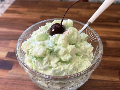 Light green fluffy salad in textured glass bowl with dark cherry on stem and white spoon