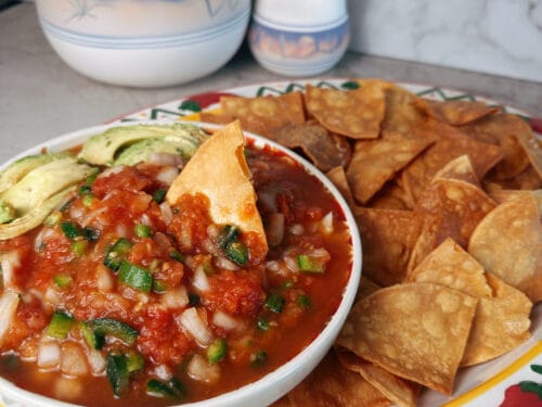 Red sauce with diced tomatoes and green onions in bowl with fried tortilla chips and avocado slices