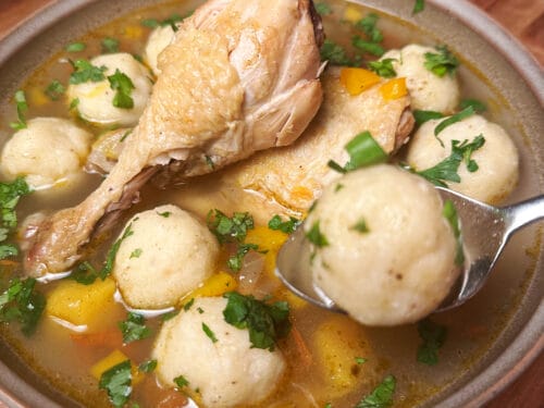 Ceramic bowl of soup with cooked chicken leg, pale dumplings, yellow pepper, and cilantro in brown broth