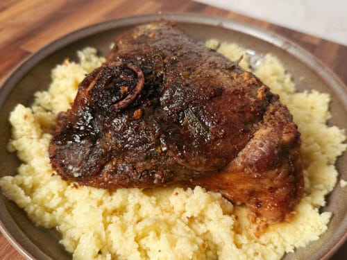 Seared meat portion with dark crust on bed of coarse yellow grain in earthenware bowl