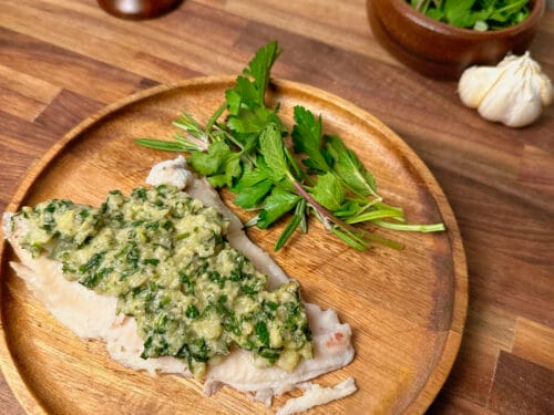 Pale fish fillet topped with green herb sauce and fresh herbs on wooden plate
