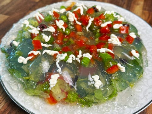 Clear gelatin mold with celery, red peppers, peas, and mayonnaise swirls on ornate plate