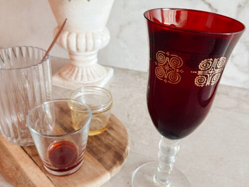 Deep red liquid in decorated goblet and small glass on marble surface with white urn holding green leaves