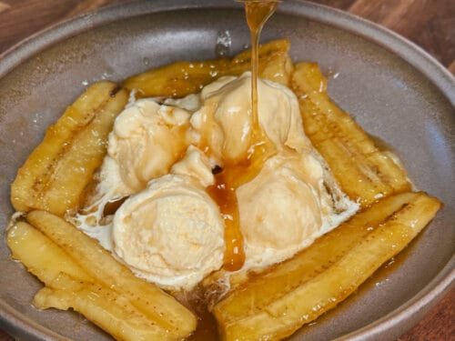 Amber sauce dripping from pan onto sautéed banana halves with vanilla ice cream scoops