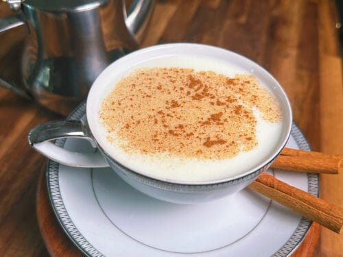 Creamy white beverage sprinkled with cinnamon, served in a bowl on a saucer with cinnamon stick, metal teapot behind
