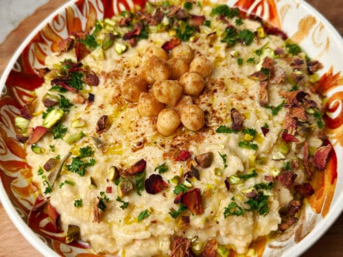 Pale beige spread garnished with chickpeas, spiced oil, pistachios, and fresh parsley on patterned plate
