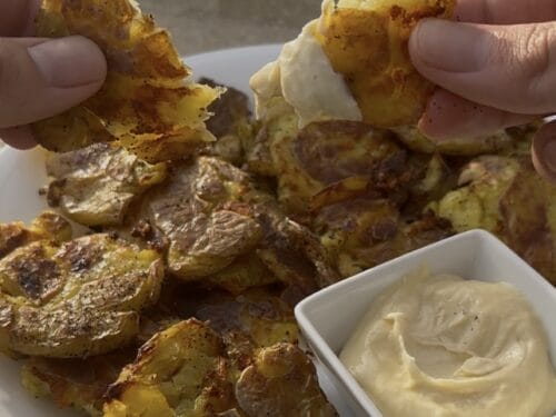 Hands holding roasted potato wedges with creamy sauce, olive oil bottles in background