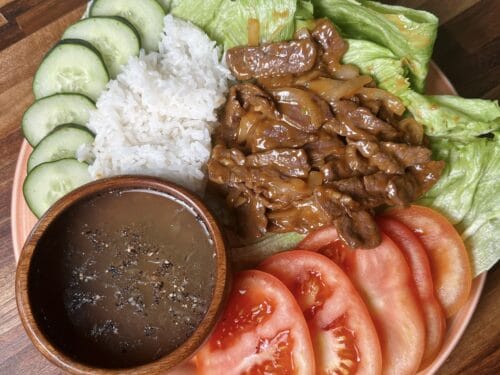 Cooked beef strips on pink plate with cucumber slices, rice, lettuce, tomato wedges, and dark sauce in wooden bowl