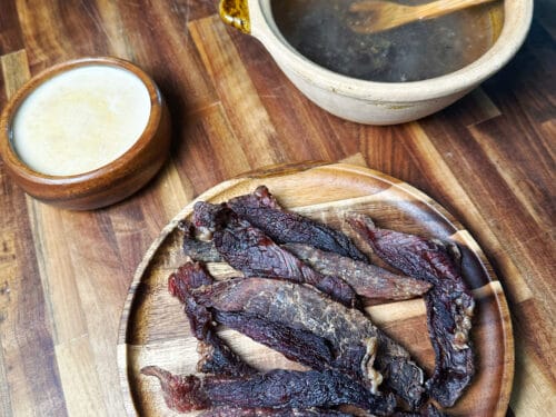 Dark dried meat strips on wooden plate with cream cup and broth bowl beside it
