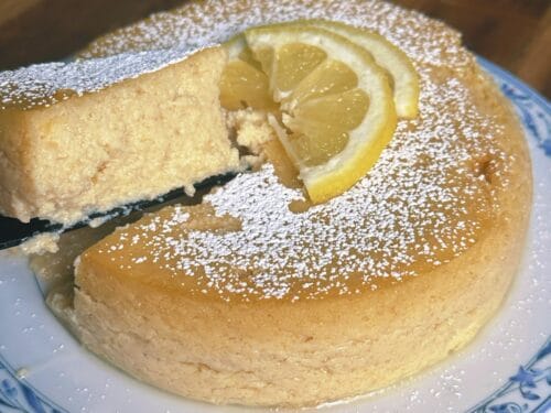 Slice of pale cake on server showing creamy interior, topped with lemon wheel and powdered sugar
