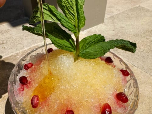 Golden syrup being poured into granita-textured drink topped with fresh mint sprigs and red seeds