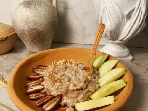 Wooden bowl of grain porridge with apple slices and green fruit, wooden spoon, stone jug, and bust sculpture