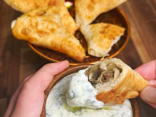 Hand holding a fried pastry cone dipped in creamy white sauce with visible meat filling inside