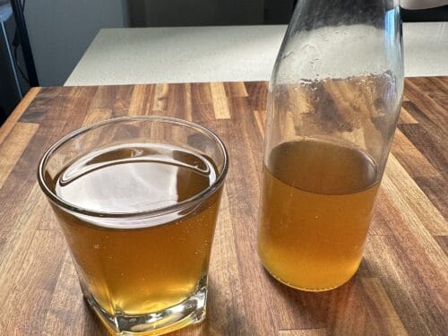 Glass of golden-brown liquid with foam head beside a filled bottle on wooden surface