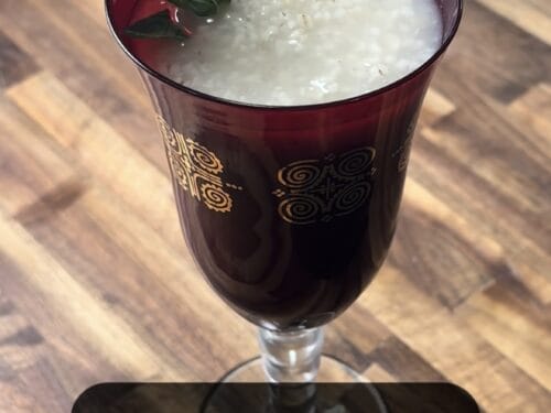 Kykeon, a cream-colored drink in a burgundy footed glass with mint sprig, on wooden surface
