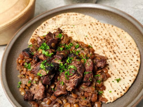 Bowl of braised lamb and lentils garnished with parsley, served on flatbread beside a wooden-lidded vessel