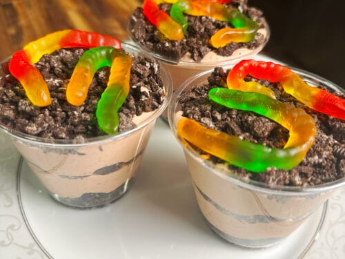 Three clear cups filled with layered chocolate pudding and crushed cookies, topped with colorful gummy worms
