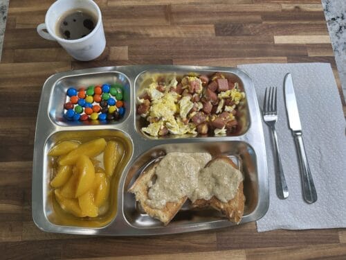 Compartmented metal tray holding colorful candies, scrambled eggs with meat, yellow peppers, and spread on toast, with coffee mug nearby