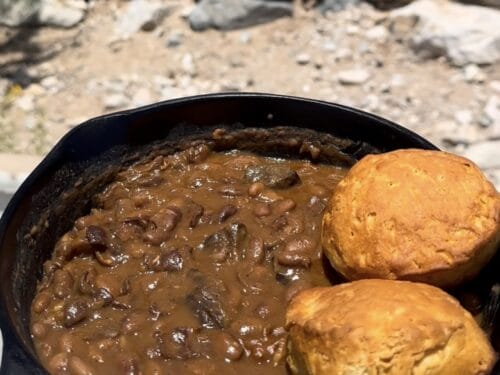 Cast-iron pot of stewed beef and beans with two golden biscuits resting on the rim, set on rocky ground