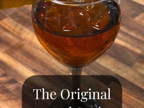 Hand holding orange wedge above glass of reddish-brown liquid with ice on wooden surface