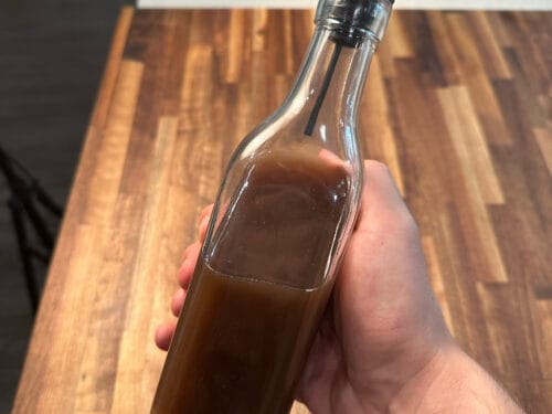 Hand holding glass bottle with metal pour spout filled with reddish-brown liquid